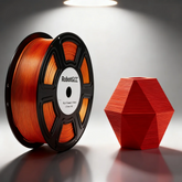 Spool of orange 3D printing filament and a red 3D printed vase on a reflective surface.
